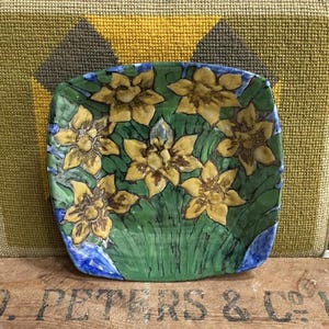 May include: Square ceramic plate with a hand-painted floral design. The plate features yellow flowers with brown details on a green background with blue accents. The plate is set against a textured background.