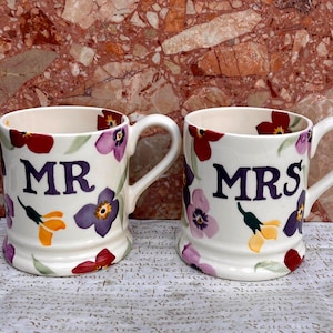 May include: Two cream-colored ceramic mugs featuring a floral pattern with red, purple, and yellow flowers. The left mug has the text MR printed in dark purple, and the right mug has the text MRS printed in dark purple. Both mugs have a handle.