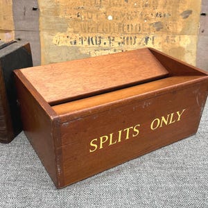 Op de afbeelding: Een rechthoekige houten kist met een schuin deksel en een medium bruine afwerking. Het voorpaneel is voorzien van de tekst SPLITS ONLY in gele hoofdletters. De kist heeft een zichtbare houtnerf en lichte slijtage aan de randen.