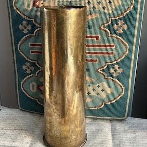 Puede incluir: Un jarrón alto de color latón con diseños grabados de figuras y patrones. El jarrón tiene forma cilíndrica con una base ligeramente ensanchada. Se encuentra sobre una superficie con texto manuscrito y un fondo estampado en verde azulado y beige.