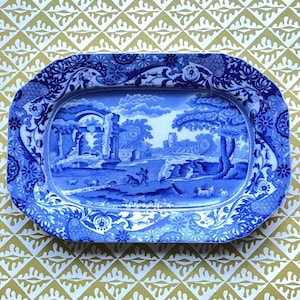 Può includere: Un piatto da portata ovale in ceramica con un motivo a trasferimento blu e bianco. Il centro raffigura un paesaggio pastorale con rovine, alberi e figure, mentre l'ampio bordo presenta un dettagliato disegno floreale e a volute in blu.