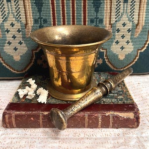Pode incluir: Um conjunto de almofariz e pilão de latão com um almofariz de latão polido de borda larga e um pilão de latão texturizado a condizer. O conjunto está colocado sobre um livro antigo encadernado em pele com lombada vermelha escura e capa estampada.