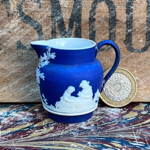 May include: A small blue and white ceramic jug with a white cameo design of two women. The jug has a handle and a spout. The jug is approximately 5 cm tall.