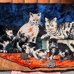 May include: A rectangular wall hanging featuring a woven scene of five cats in a room with a fireplace. The cats are depicted in black, white, and gray tones on a vibrant orange rug against a deep blue background, finished with blue fringe.