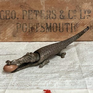 May include: A vintage metal alligator nutcracker with a nut in its jaws. The nutcracker is bronze-colored and detailed with scales and textured features. The background includes a wooden sign with text.