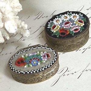 May include: Two oval metal trinket boxes with hinged lids featuring intricate micro-mosaic floral designs. One box has a black background with white, red, and pink flowers, while the other has a white background with red, blue, and green floral patterns.