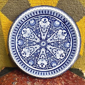 Puede incluir: Un plato decorativo redondo, azul y blanco, con un intrincado diseño floral y geométrico. El plato presenta un borde detallado y un motivo floral central. El plato está colocado sobre una superficie estampada.