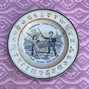 May include: A round ceramic plate with a cream-coloured centre and a dark rim. The centre features a black and white illustration of two children, labelled JUVENILE COMPANIONS and THE YOUNG SERGEANT. The alphabet is embossed around the border.