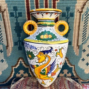 May include: A hand-painted ceramic vase with a yellow, orange, blue, and green design. The vase features a mythical creature and floral motifs. It has two ring handles and measures approximately 7 inches in height.