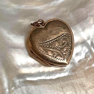 May include: A heart-shaped gold locket with an engraved design. The locket is closed and has a small loop at the top for a chain. The background is a textured, off-white surface. The locket is likely made of gold.