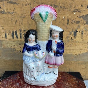 Könnte beinhalten: Vintage Porzellanfigur mit zwei Kindern und einem dekorativen Blumenelement. Die Kinder tragen blaue, weiße und rosa Kleidung. Die Figur hat einen cremefarbenen Sockel und ein strukturiertes, korbartiges Design mit einem rosa und grünen Oberteil.