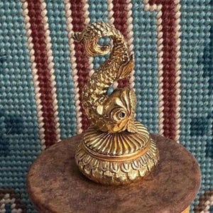 May include: A gold-toned metal trinket box featuring a sculpted fish figure on the lid. The fish has detailed scales and a curved tail. The base of the box is circular with a ribbed design, and the entire object has a polished, reflective finish.