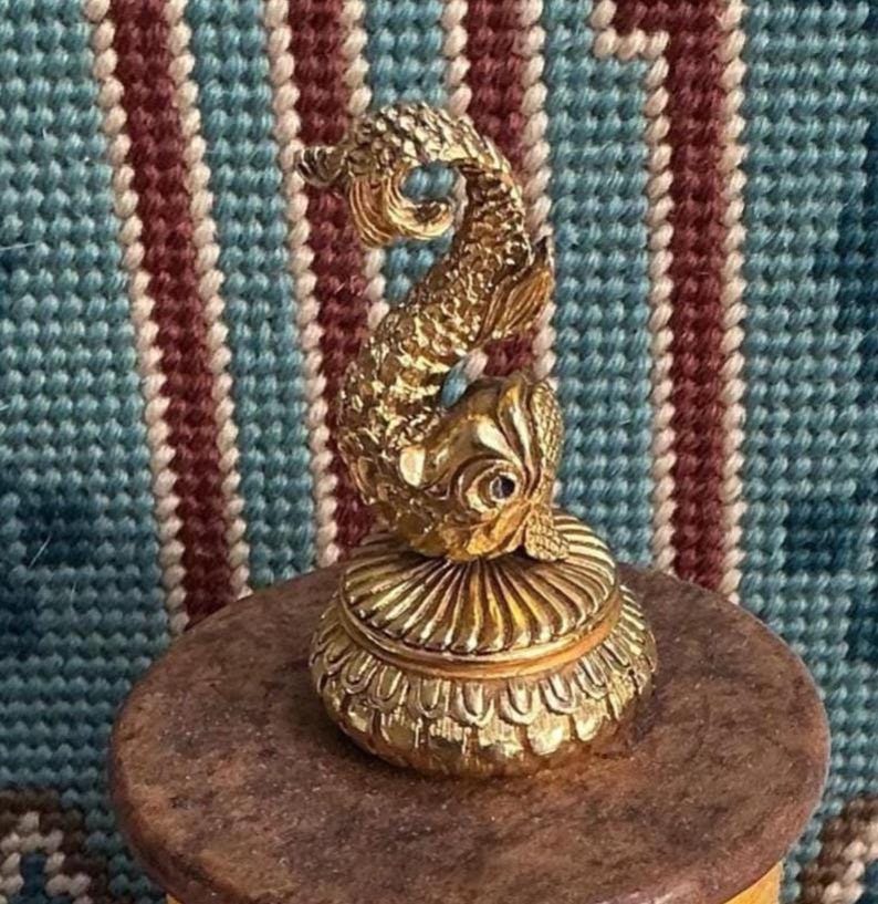 May include: A gold-toned metal trinket box featuring a sculpted fish figure on the lid. The fish has detailed scales and a curved tail. The base of the box is circular with a ribbed design, and the entire object has a polished, reflective finish.
