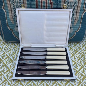 May include: A set of six stainless steel knives with cream-coloured handles, presented in a white box with a black velvet interior. The box is open, revealing the knives neatly arranged. The handles are a light cream colour.
