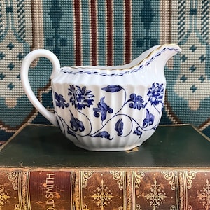 May include: White porcelain creamer with blue floral pattern. The creamer has a curved handle and a spout with gold trim. The blue floral design wraps around the body. The creamer is on a vintage book.