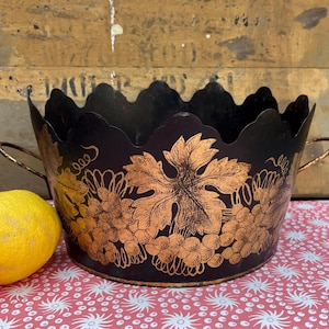 May include: A black metal wine bucket with gold grape leaf and vine detailing. The bucket has a scalloped rim and two small handles. A yellow lemon sits to the left of the bucket on a red and white patterned surface.