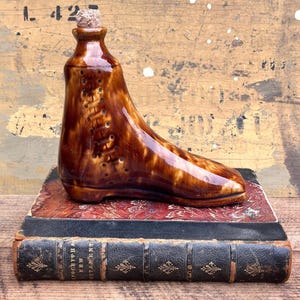 May include: A brown ceramic boot-shaped bottle with a cork stopper. The boot has a glossy finish and is placed on top of two antique books with decorative covers. The boot has a lace-up design.