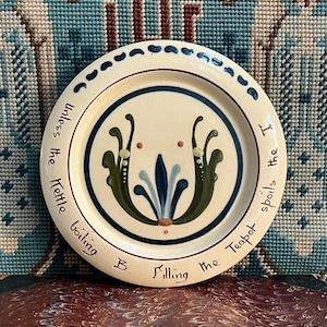 Puede incluir: Un plato de cerámica color crema con un diseño floral azul y verde en el centro. El plato tiene un borde azul y una cita alrededor del borde que dice: "Unless the kettle boiling B Filling the Teapot spoils the I."