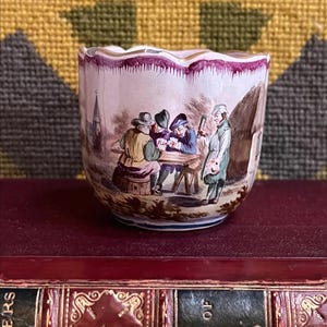 May include: A small, antique porcelain cup with a scalloped rim. The cup features a detailed, hand-painted scene of several figures gathered around a table, with a church and thatched-roof building in the background. The cup is set on a red book.