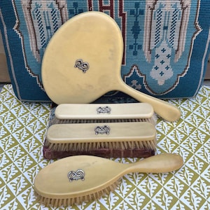 May include: A vintage vanity set featuring a hand mirror and three brushes. The set is a creamy yellow color with silver-toned embellishments. The brushes have natural bristles. The background includes a teal tapestry with angel figures and a patterned green and white surface.