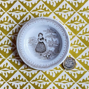 May include: A small white ceramic plate featuring a black transfer print of a person standing in a field with a lamb and trees. The rim has a decorative dotted pattern. A one-pound coin is placed next to the plate for scale, showing the size.