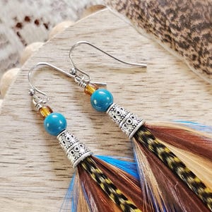 May include: A pair of dangle earrings featuring a silver-tone metal cone with a decorative pattern, turquoise beads, and colorful feathers in shades of brown, blue, and yellow. The earrings have a hook closure.