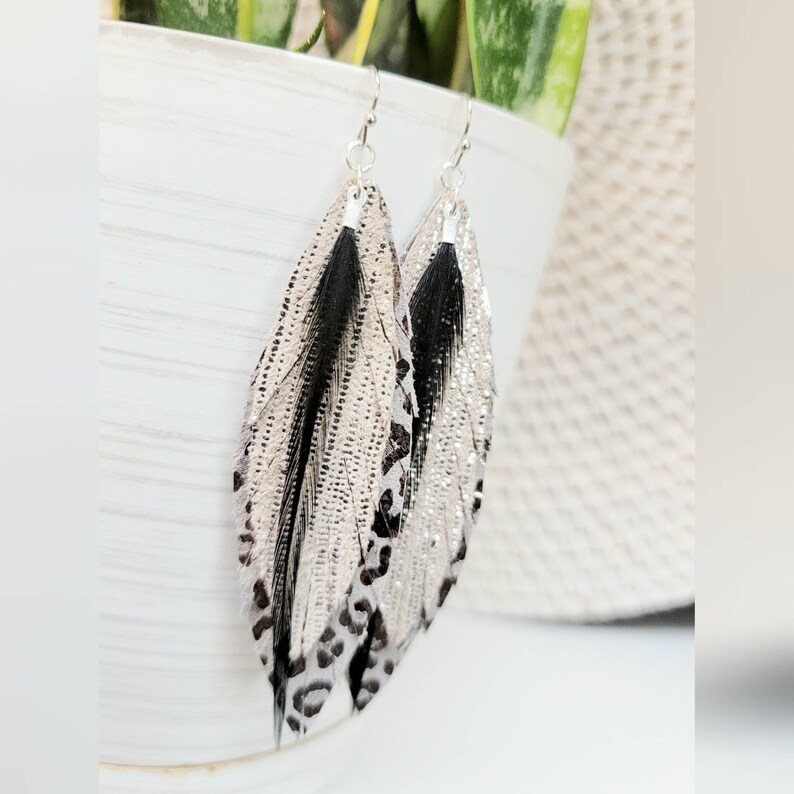 May include: A pair of dangle earrings featuring a layered feather design with black, white, and silver accents. The feathers have a leopard print pattern.