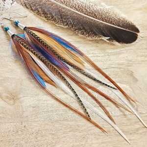 May include: A pair of long feather earrings with brown, blue, and white feathers. The earrings have silver hooks and blue beads. A large feather rests nearby on a wooden surface. The earrings are a bohemian style.