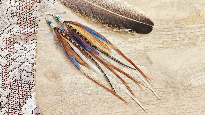 May include: A pair of long feather earrings with brown, blue, and white feathers. The earrings have silver hooks and blue beads. A large feather rests nearby on a wooden surface. The earrings are a bohemian style.