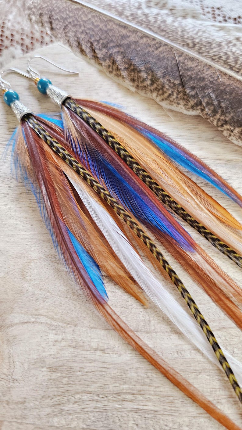 May include: Long feather earrings with a silver accent and teal bead. The earrings feature a mix of brown, blue, white, and orange feathers. The feathers are arranged in a layered design, creating a unique and eye-catching accessory.