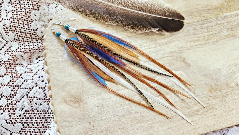 May include: A pair of long feather earrings with silver hooks and blue beads. The earrings feature brown, white, and blue feathers. A large feather rests in the background on a wooden surface.