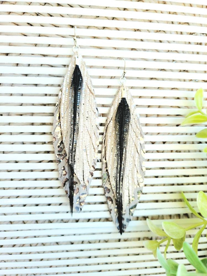 May include: A pair of long, silver feather earrings with black feathers. The earrings are made of leather and have a metallic finish.
