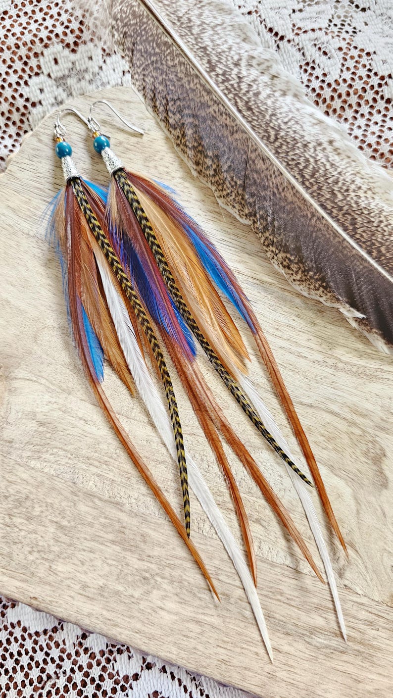 May include: Long feather earrings with silver hooks and teal beads. The earrings feature a mix of brown, white, blue, and gold feathers. A large brown and gray feather lies in the background. The earrings are on a wooden surface.