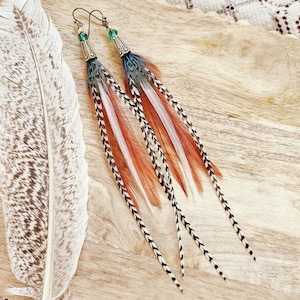 May include: Long feather earrings with a mix of colors including red, white, black, and blue. The earrings have a metal top with a green bead and a hook closure. The feathers are arranged in a layered design, creating a unique and eye-catching accessory.