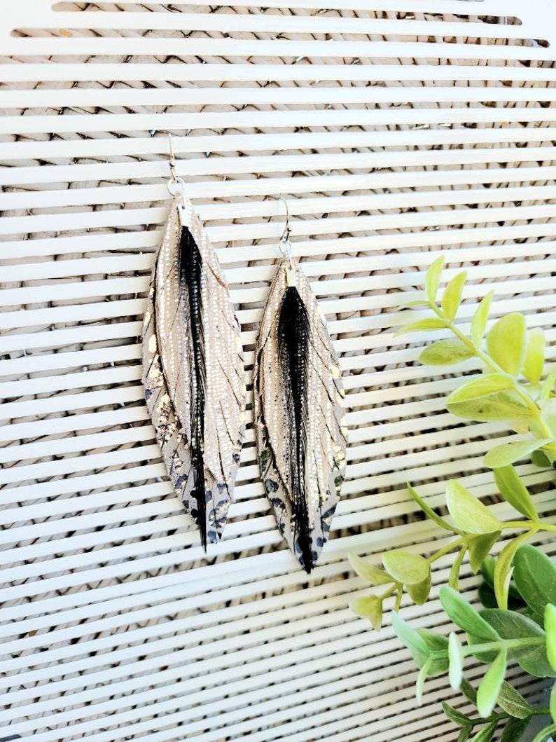 May include: A pair of long, silver and black feather earrings. The earrings are made of leather and have a metallic finish. The feathers are black and have a natural look.