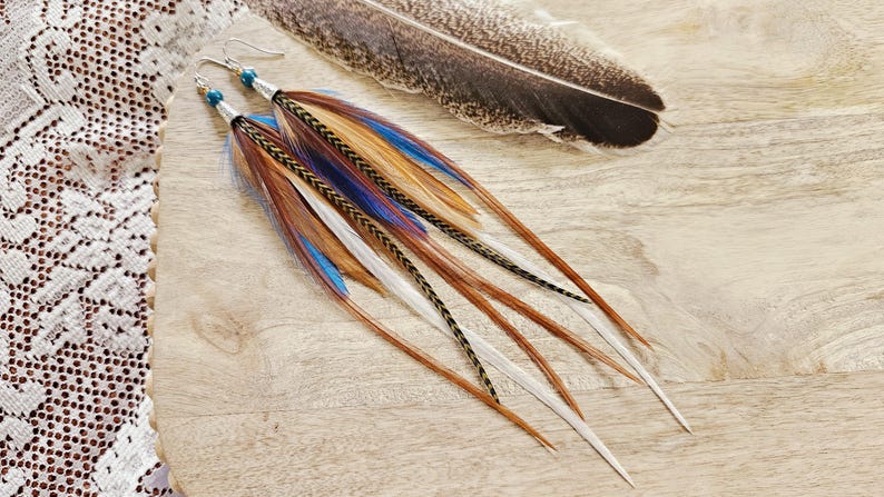 May include: Long feather earrings with brown, blue, and white feathers. The earrings have silver hooks and small blue beads. A large feather rests nearby on a wooden surface.