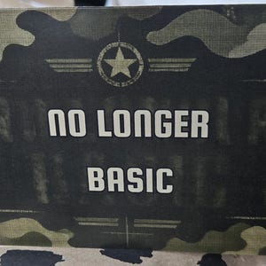 May include: A rectangular card featuring a green, brown, and black camouflage pattern. Centered at the top is a circular emblem with a star. The text "NO LONGER" appears above the word "BASIC" in a bold, white, sans-serif font on a dark background.