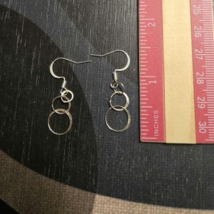May include: A pair of silver-toned dangle earrings featuring three interlocking circular rings of varying sizes suspended from a standard hook wire. A ruler is positioned next to the earrings, showing a total length of approximately 1.25 inches.