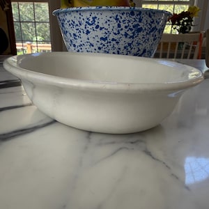 May include: A plain white oval ceramic basin with a wide rim, positioned in the foreground. Behind it sits a large, deep, round bowl featuring a white base with a speckled blue enamel pattern, containing several yellow bananas and a mango.