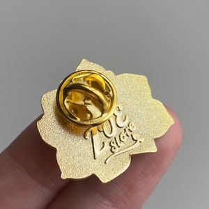 Puede incluir: Pin de esmalte de color dorado con un dise&ntilde;o floral. La parte posterior del pin tiene un cierre de seguridad y el texto "Love Store" en una fuente cursiva.