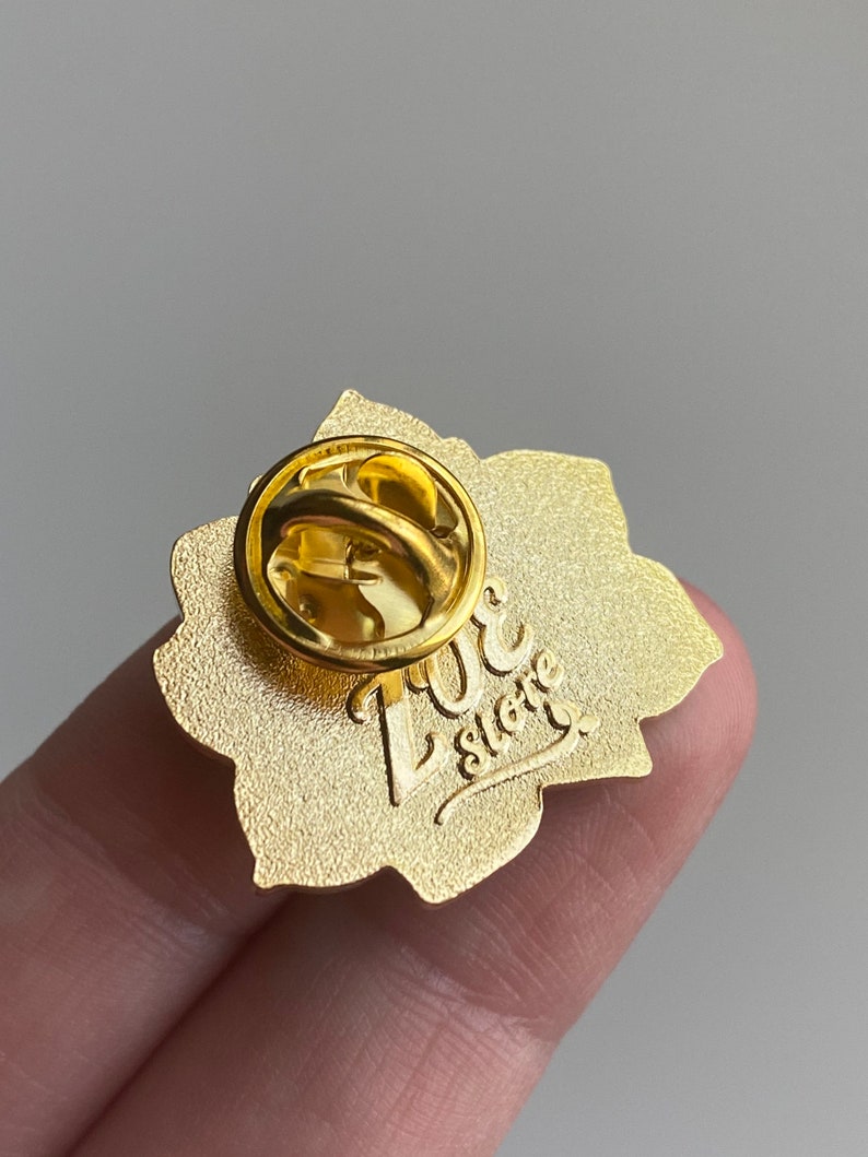 Puede incluir: Pin de esmalte de color dorado con un dise&ntilde;o floral. La parte posterior del pin tiene un cierre de seguridad y el texto "Love Store" en una fuente cursiva.