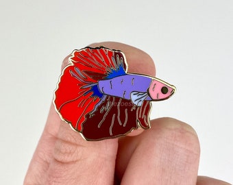 May include: A gold-tone enamel pin featuring a red, purple, and blue betta fish with a pink mouth. The fish is depicted with its fins spread out, creating a flowing, elegant design.