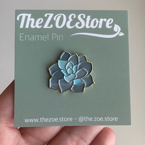 Puede incluir: Un pin de esmalte azul y verde con una planta suculenta. El pin est&aacute; en una tarjeta verde con el texto "The ZOEStore Enamel Pin" y "www.thezoe.store - @the.zoe.store".