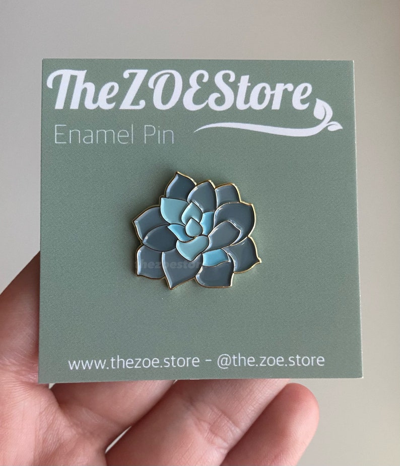 Puede incluir: Un pin de esmalte azul y verde con una planta suculenta. El pin est&aacute; en una tarjeta verde con el texto "The ZOEStore Enamel Pin" y "www.thezoe.store - @the.zoe.store".