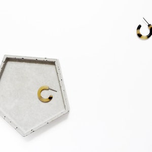 May include: Two pairs of hoop earrings on a white background. One pair is in a concrete tray, the other pair is on the surface. Both pairs are black and brown.