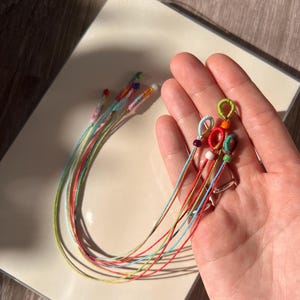Puede incluir: Una colección de pulseras coloridas hechas a mano con extremos en bucle y detalles de cuentas. Las pulseras presentan una variedad de colores brillantes, incluyendo rojo, azul, verde y amarillo. Las pulseras se muestran sobre una superficie blanca.