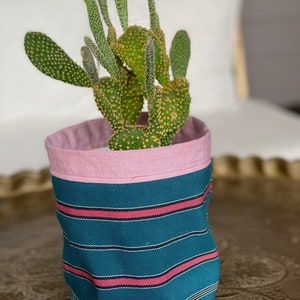 May include: A green cactus with yellow spots is planted in a teal fabric pot with pink and red stripes. The pot has a pink interior and sits on a decorative gold surface. The cactus is a houseplant.
