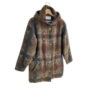 May include: A hooded jacket featuring a multi-coloured geometric pattern in shades of brown, green, and burgundy. The jacket has a button-front closure, long sleeves, and two front pockets. An L.L. Bean label is visible on the inner collar.