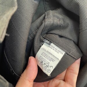 以下が含まれることがあります： 文字M、Made in Japan、100% Polyesterと記載された白い内側タグが付いたダークグレーの衣類。タグには、ドライクリーニング、タンブル乾燥禁止、低温アイロン掛けを示すケアシンボルが記載されています。