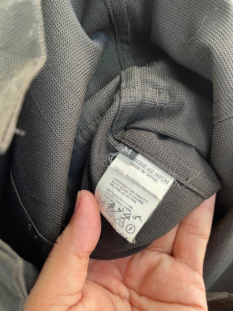 以下が含まれることがあります： 文字M、Made in Japan、100% Polyesterと記載された白い内側タグが付いたダークグレーの衣類。タグには、ドライクリーニング、タンブル乾燥禁止、低温アイロン掛けを示すケアシンボルが記載されています。